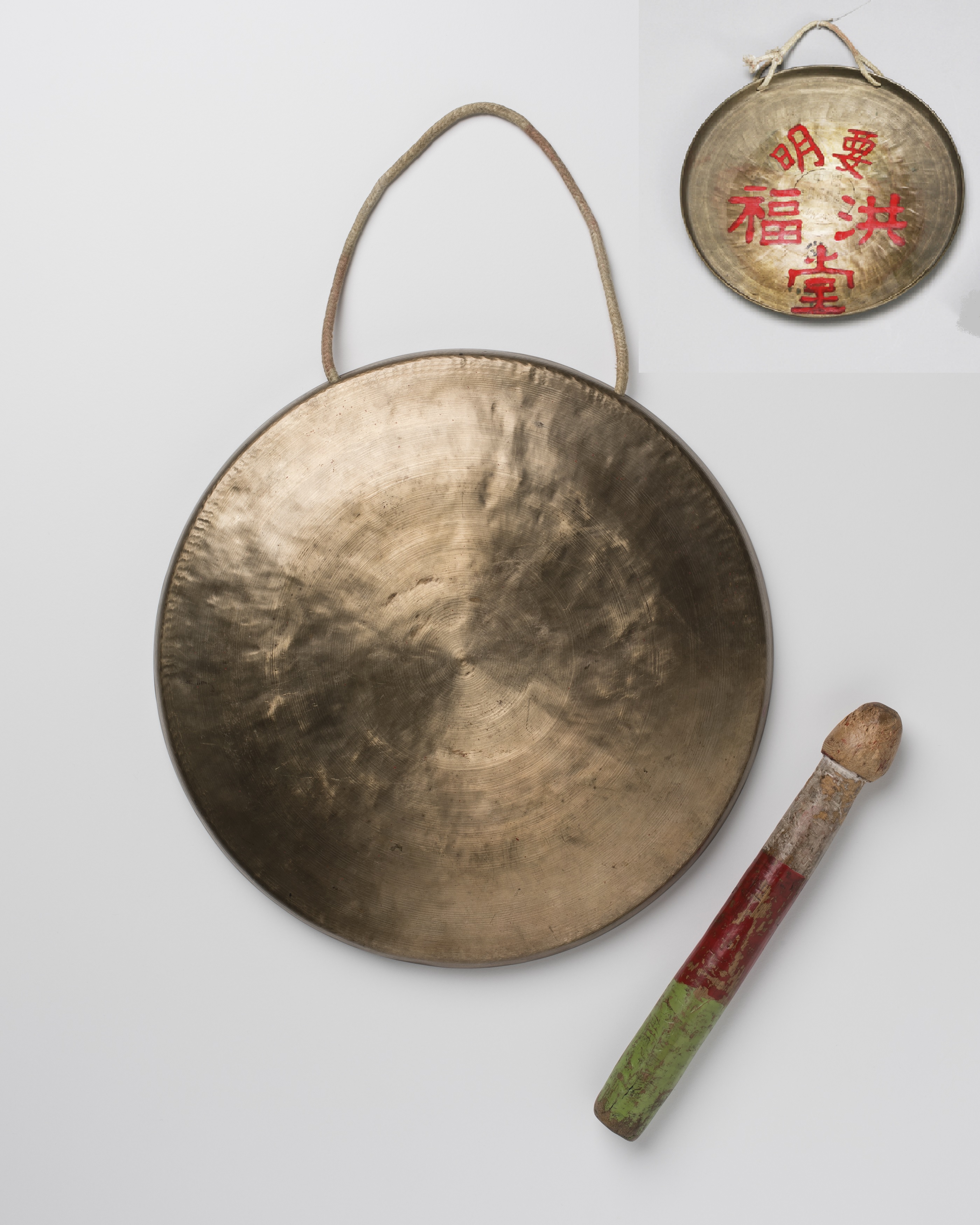 An image of a circular brass gong with cotton cord handle attached with its painted wooden beater to the side. A small image of the reverse of the gong with Chinese characters in red is in the top right corner of the image.